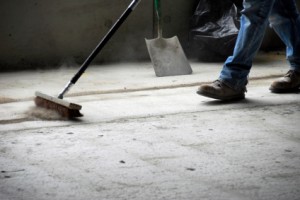 The legs and feet of a construction worker sweeping up on rough concrete at a job site using a large broom